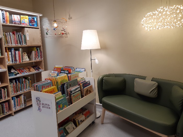 Rum med beige vägg, träfärgad bokhylla med böcker, vitt bilderbokstråg, grön soffa samt vit golvlampa och två hängande taklampor. 