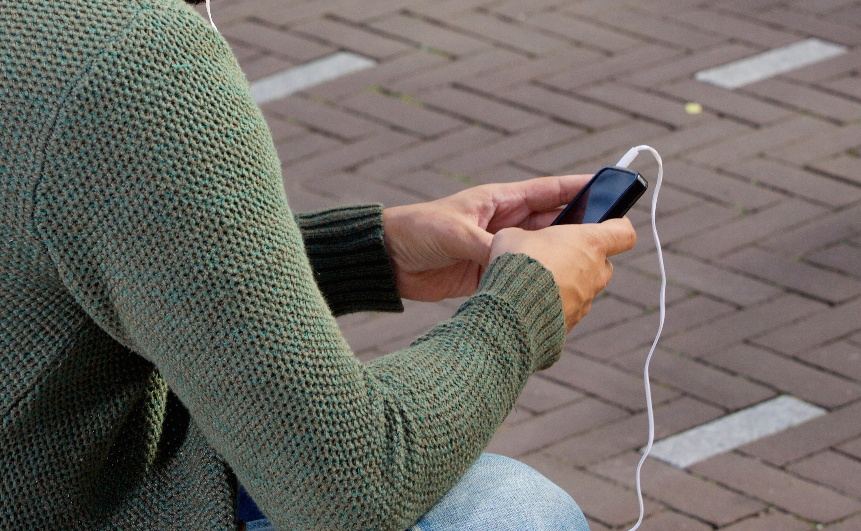 Närbild på person i grön stickad tröja som sitter utomhus vid stenlagd gata/torg, tittandes i mobiltelefon med inkopplade hörlurar.